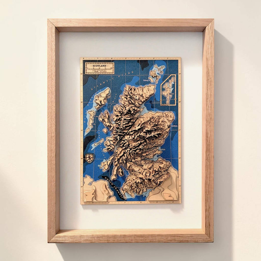 A framed 3D wooden topographic map of Scotland, showcasing raised mountain peaks in natural wood tones over a deep blue ocean background, housed in a light oak frame and mounted on a neutral wall.