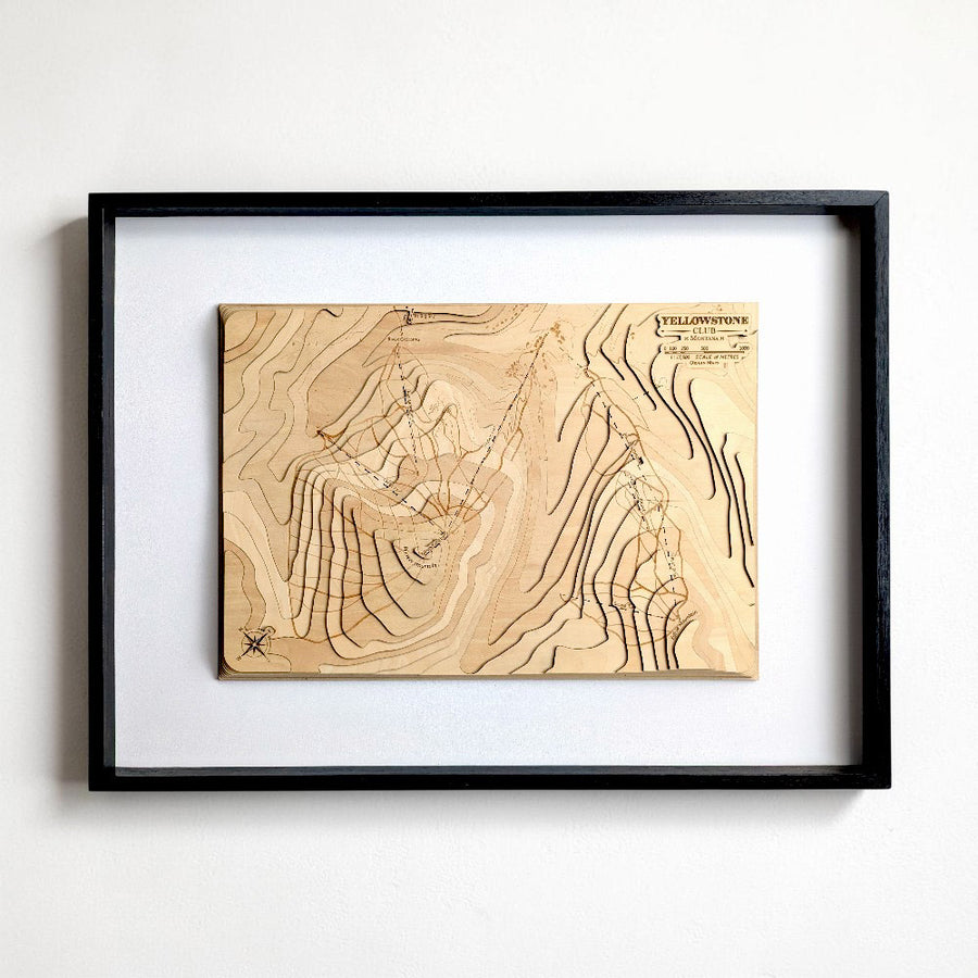 A framed wooden topographic map of Yellowstone club ski resort in Montana, featuring detailed contour lines and engraved text, mounted on a white mat inside a black frame against a white wall.
