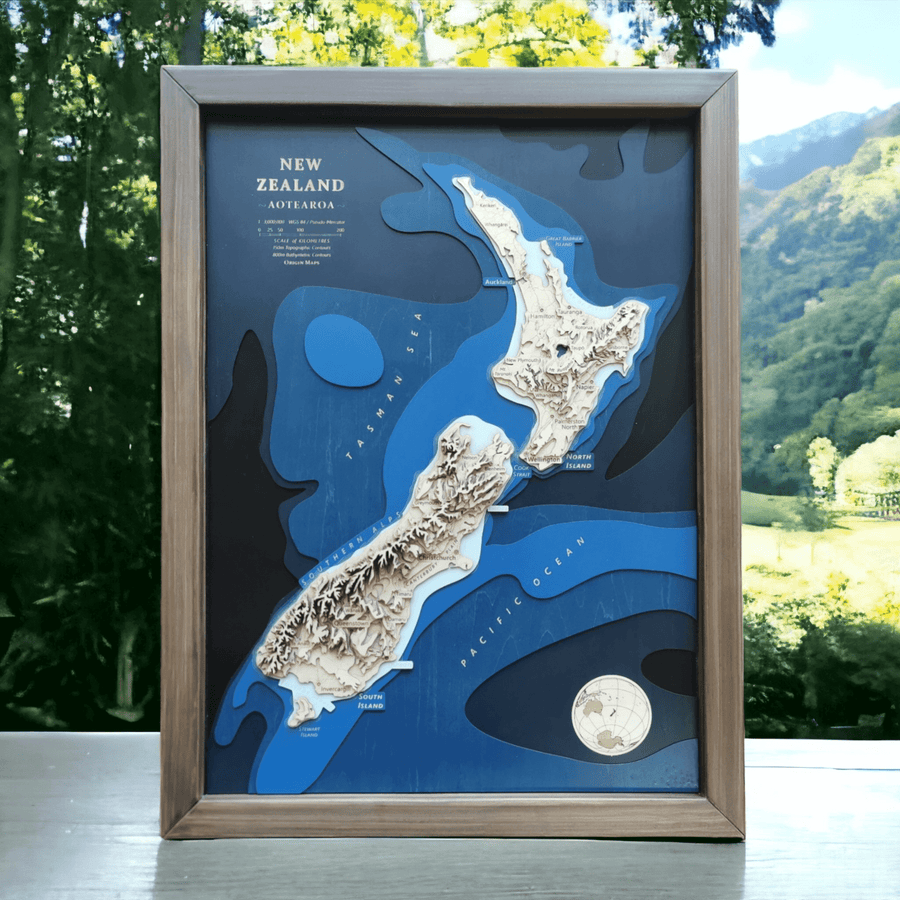 Framed wooden topographic contour map of new zealand featuring the bathymetry and the topography through contours. It is sitting on a bench with a scenic new zealand landscape in the background