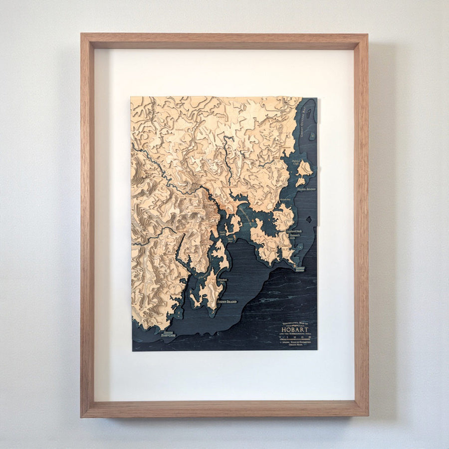 A framed wooden topographical map print of Hobart, Tasmania, featuring light beige land and dark navy blue water, housed in a light oak wood frame with a white mat, mounted on a neutral gray wall.