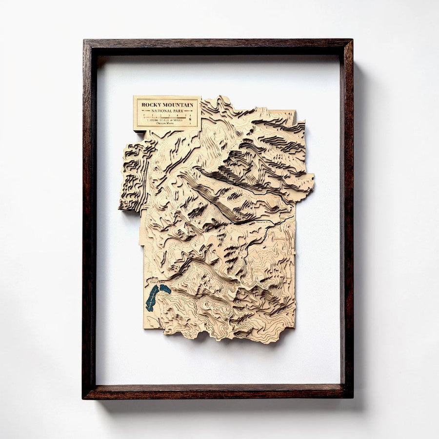 A framed 3D laser-cut wooden topographic map of Rocky Mountain National Park, Colorado, featuring a dark brown wood frame and a cream-colored label, mounted on a white wall.